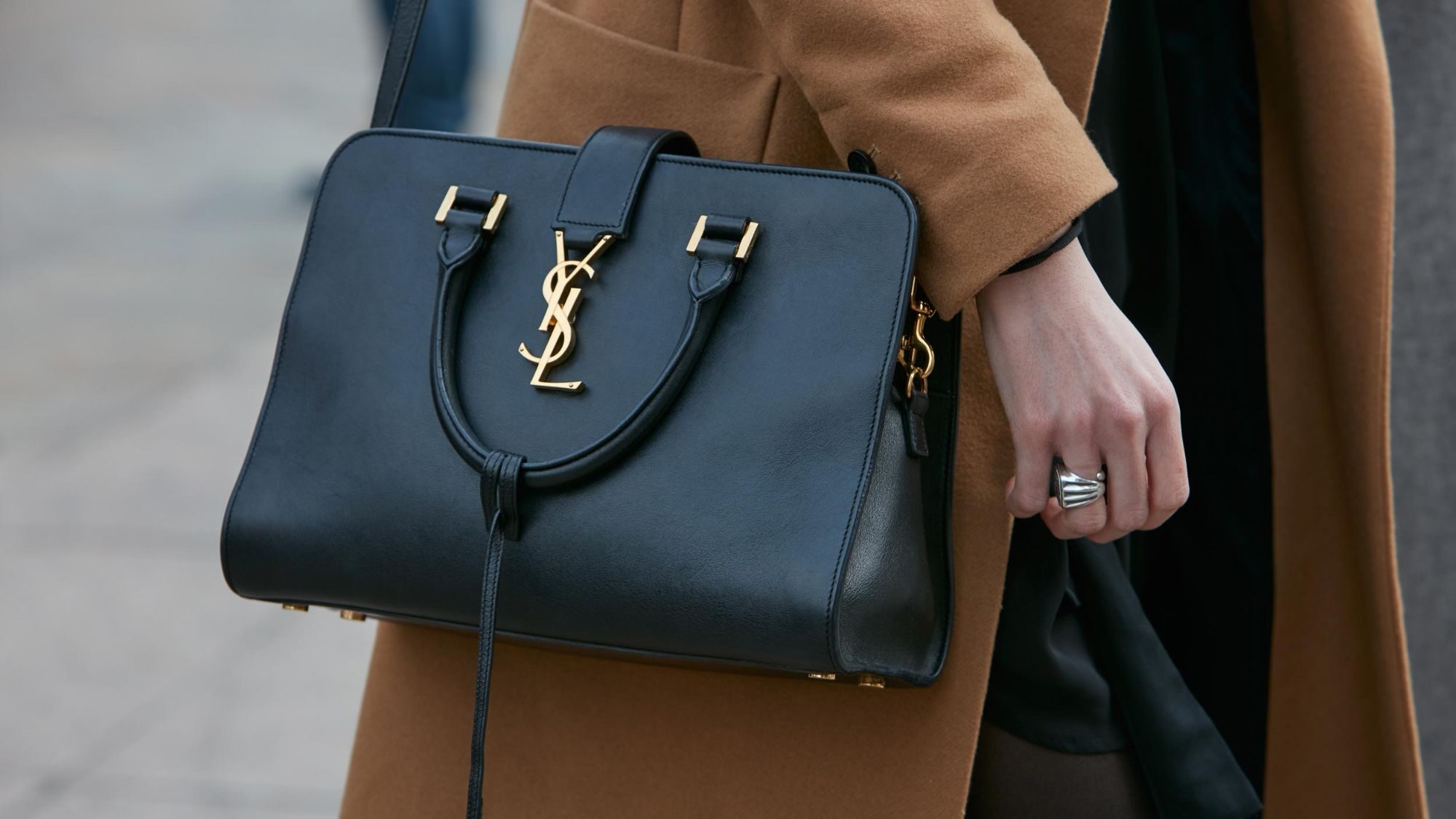 Black leather handbag with a gold YSL logo, carried by a person in a camel coat.