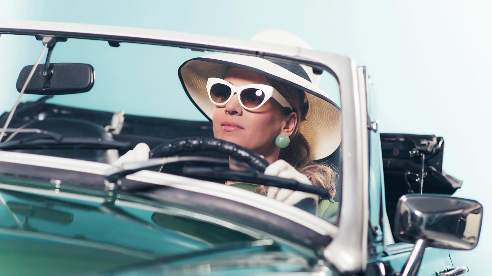 A woman in a wide sun hat, cat-eye sunglasses and white gloves drives a turquoise convertible.